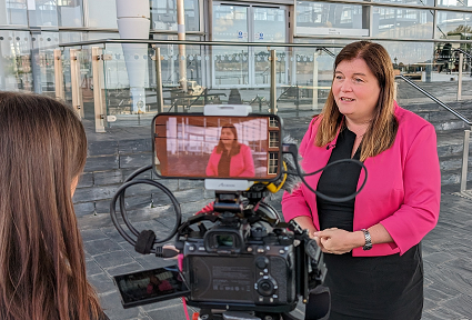 Mae Sioned Williams AS yn cael ei chyfweld ar gyfer y teledu a gellir gweld camera yn y blaendir, gyda sgrin fach yn dangos y llun o Sioned. Mae Sioned Williams MS yn sefyll ar risiau'r Senedd, y tu allan i adeilad y Senedd, y mae ei blaen gwydr i'w weld y tu ôl iddi.