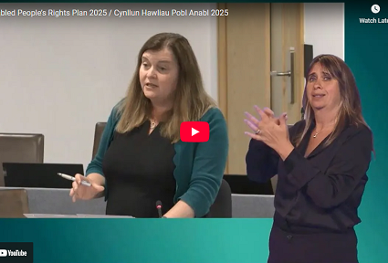 Sioned Williams AS yn siarad yn Siambr y Senedd yn ystod dadl ar Gynllun Hawliau Pobl Anabl 2025. Mae’n dal pin ac yn sefyll wrth ddesg. Mae cyfieithydd Iaith Arwyddion Prydain (BSL) i’w weld ar ochr dde’r sgrin.