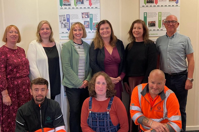 Mae grŵp o wyth o bobl yn sefyll am lun y tu mewn i swyddfa neu adeilad sefydliad cymunedol. Mae pump o bobl yn sefyll mewn rhes yn y cefn, tra bod tri yn penlinio yn y blaen. Mae dau aelod o'r grŵp yn gwisgo dillad gwaith amlwg, ac mae byrddau arddangos hanesyddol o'r enw 