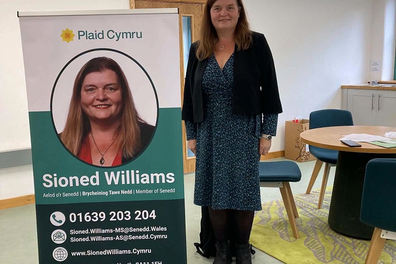 Mae Sioned yn sefyll wrth ymyl baner rolio Plaid Cymru mewn ystafell gyfarfod olau. Mae’r faner yn cynnwys portread, manylion cyswllt, cyfeiriad gwefan a gwybodaeth am leoliad swyddfa. Mae byrddau crwn a chadeiriau glas i’w gweld yn yr ystafell, gyda phapurau ar un o’r byrddau a drws pren yn y cefndir.