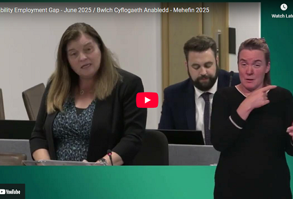 Aelod o’r Senedd, Sioned Williams, yn siarad yn Siambr y Senedd yn ystod dadl ar y bwlch cyflogaeth i bobl anabl ym Mehefin 2025. Mae dyn yn eistedd y tu ôl iddi, ac mae cyfieithydd Iaith Arwyddion Prydain (BSL) ar ochr dde’r sgrin