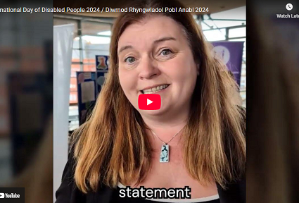 Mae Sioned Williams AS yn cyflwyno neges fideo ar gyfer Diwrnod Rhyngwladol Pobl Anabl 2024. Mae hi’n gwenu ac yn siarad yn uniongyrchol â’r camera, gan sefyll y tu mewn i’r Senedd gydag arwyddion i’w gweld yn y cefndir