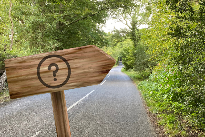 Arwydd ffordd gyda marc cwestiwn arno yn pwyntio i lawr lôn wledig ddi-nodwedd