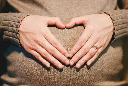 A lady touching the pregnant belly with two hands making the shape of a heart