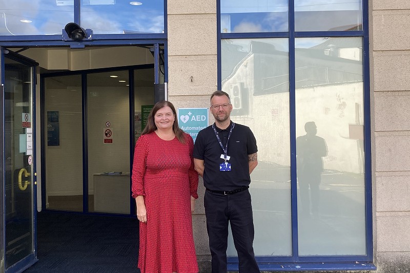 Mae dau berson yn sefyll y tu allan i fynedfa swyddfa Canolfan Byd Gwaith o dan arwydd gwyrdd dwyieithog yn darllen “canolfan byd gwaith canolfan byd gwaith.” Mae gan yr adeilad ffenestri mawr â ffrâm las a drws agored. Mae arwydd glas sy'n nodi lleoliad diffibriliwr allanol awtomataidd (AED) wedi'i osod wrth ymyl y fynedfa, ac mae ardal gerddwyr wedi'i phalmantu â brics i'w gweld yn y blaendir.