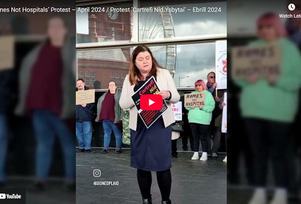 Sioned Williams yn siarad mewn protest y tu allan i’r Senedd gan ddal arwydd sy’n darllen ‘Cartrefi Nid Ysbytai’, gyda chefnogwyr yn sefyll y tu ôl iddi’n dal placardiau