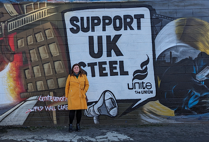 Sioned William yn sefyll o flaen celf wal graffiti 'Support UK Steel'