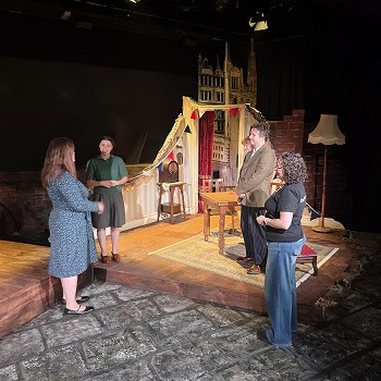 People discussing a theatre set featuring period furniture and streetscape scenery on a stage at Theatr na nÔg.