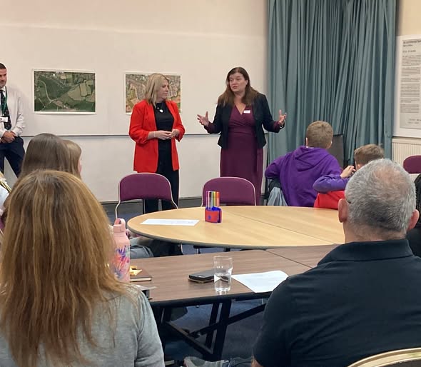 .People are seated around round tables in a meeting room while two speakers stand at the front addressing the group. One speaker wears a dark jacket over a burgundy outfit and gestures while speaking, while the other wears a bright red jacket. Maps and information boards hang on the walls behind them, and a blue curtain covers one side of the room. Papers, drinks and stationery are placed on the tables as attendees listen to the presentation.