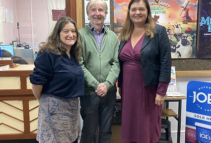 Mae Sioned a dau berson yn sefyll yng nghyntedd lleoliad cymunedol wrth ymyl bwrdd hysbysiadau sy'n arddangos posteri theatr a gwybodaeth leol. Uwchben y rhain mae arwydd dwyieithog yn darllen “Swyddfa Docynnau / Ymholiadau” a “Swyddfa Docynnau / Ymholiadau.” Mae'r bwrdd hysbysiadau yn cynnwys posteri hyrwyddo, gan gynnwys un ar gyfer Shaun the Sheep: The Beast of Mossy Bottom. Mae ardal dderbynfa i'w gweld trwy ffenestr wasanaeth yn y cefndir, tra bod stondinau a arddangosfeydd gwybodaeth gymunedol wedi'u lleoli o amgylch y cyntedd.