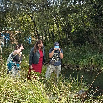Mae tri o bobl yn sefyll wrth ymyl dyfrffordd gul mewn cynefin gwlyptir wedi'i amgylchynu gan laswellt tal, cyrs a choed. Mae un person yn defnyddio camera neu ddyfais wylio i arsylwi bywyd gwyllt ger ymyl y dŵr, tra bod y lleill yn edrych ymlaen. Mae llystyfiant trwchus yn leinio'r lan, ac mae'r olygfa'n tynnu sylw at weithgarwch arsylwi natur a chadwraeth mewn amgylchedd dŵr croyw.