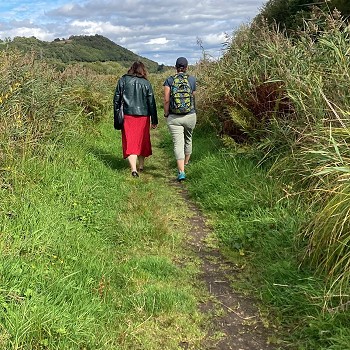 Mae dau berson yn cerdded ar hyd llwybr glaswelltog cul drwy dirwedd gwlyptir wedi'i ffinio gan gorsen tal a llystyfiant. Gellir gweld coed a bryniau isel yn y pellter o dan awyr rhannol gymylog.