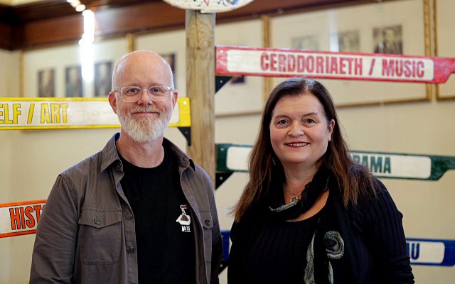 Mae dau berson yn sefyll dan arwydd Gŵyl Gelf Castell-nedd / Neath Arts Festival mewn neuadd gymunedol. Y tu ôl iddynt mae arddangosfa liwgar o arwyddion cyfeiriadol sy’n nodi gwahanol feysydd y celfyddydau, gan gynnwys Celf, Hanes, Cerddoriaeth a Drama. Mae un person yn dal llyfr, ac mae ffenestri mawr a ffotograffau mewn fframiau i’w gweld yn y cefndir.