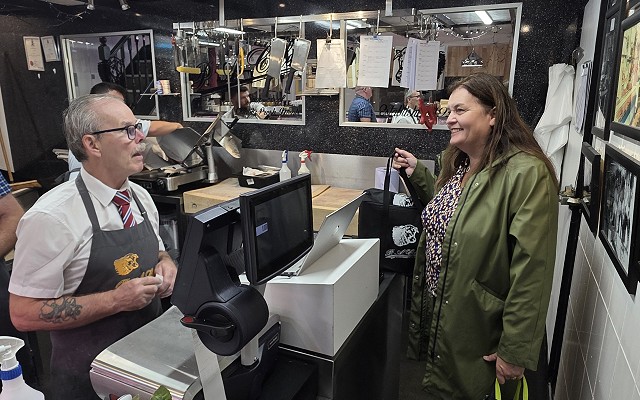 Inside a traditional butcher's shop, Sioned speaks with a member of staff standing behind the counter. The shop contains stainless-steel work surfaces, display equipment, framed photographs on the walls, and an open preparation area visible through an interior window.