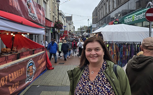 A busy town-centre street market with stalls lining both sides of a pedestrianised shopping area. Decorative bunting is strung overhead, and shoppers walk between food vendors and market stands. In the foreground, Sioned is wearing a green waterproof coat stands facing the camera, with shops including Holland & Barrett visible in the background.