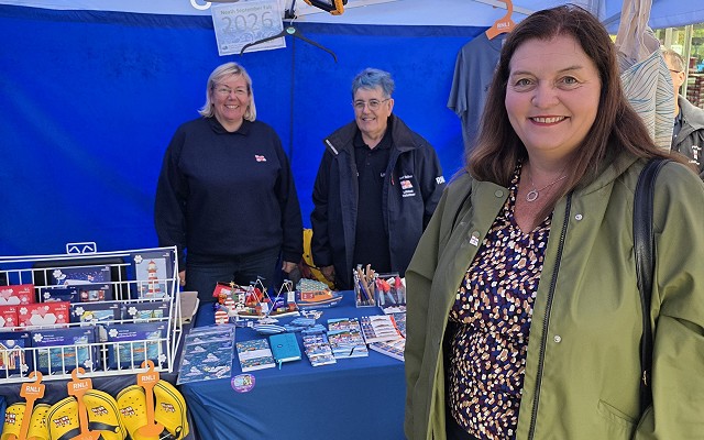 Sioned stands beside a charity stall under a blue RNLI (Royal National Lifeboat Institution) gazebo. Volunteers are positioned behind a table displaying RNLI merchandise, postcards, stationery, and fundraising items, with branded clothing and lifeboat-themed products visible throughout the stand.