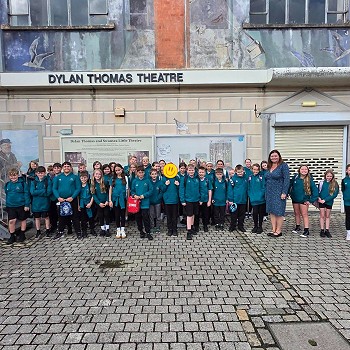 A large group of school pupils and accompanying adults stand outside the Dylan Thomas Theatre in Swansea. The theatre's name is displayed above the entrance, while a mural decorates the upper façade. The group is gathered for a photograph in the paved area outside the building.