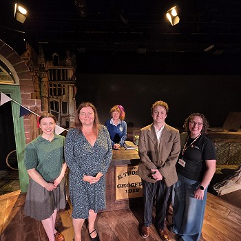 Group standing on a theatre stage in front of a period shop set during a visit to a Theatr na nÔg production.