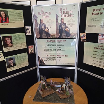 Exhibition display featuring posters for the bilingual production Ac Abertawe'n Fflam / It'll All Be Over By Christmas, cast and creative team information, and a scale model of the theatre set on a round table.